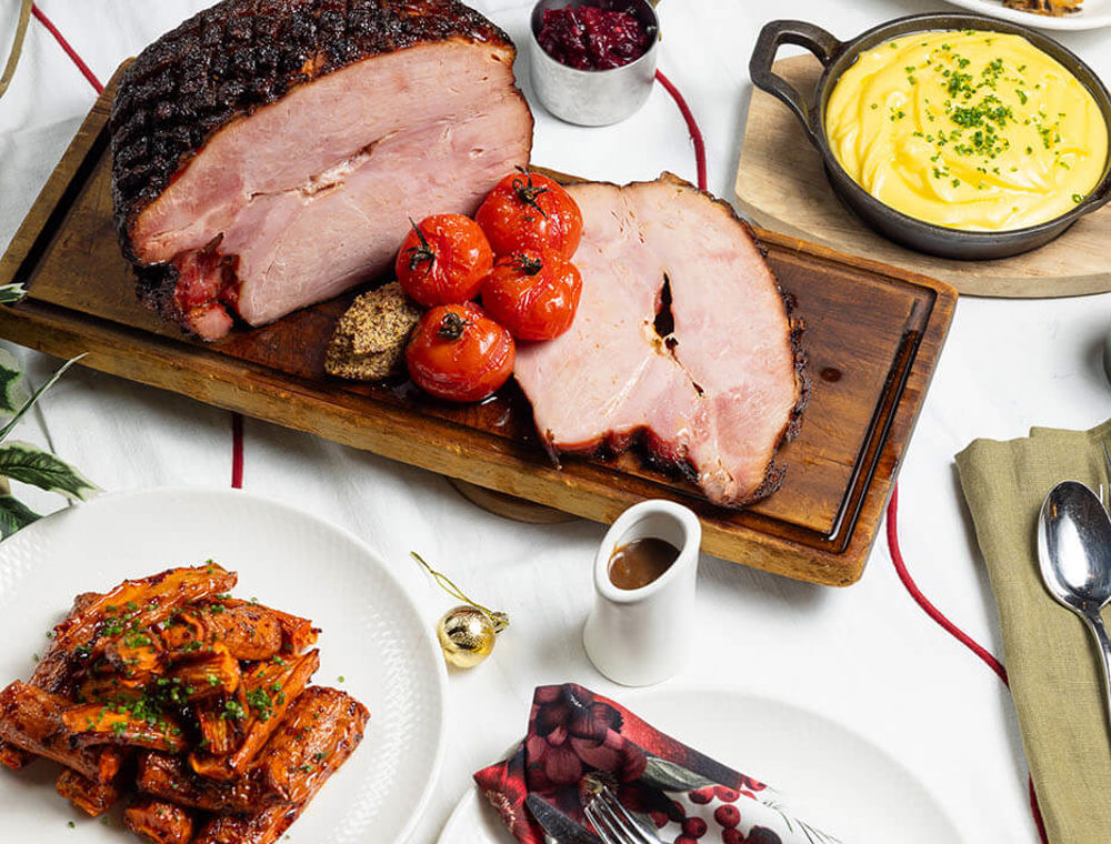 Maple glazed ham carved on a wooden board with roasted tomatoes and mashed potato.